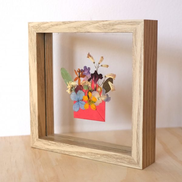 Pressed Flower Art Blossom Mail featuring flowers popping out from a mini red envelope and framed in a double glass wooden frame which is on an angle to the left and stands along on a wood surface
