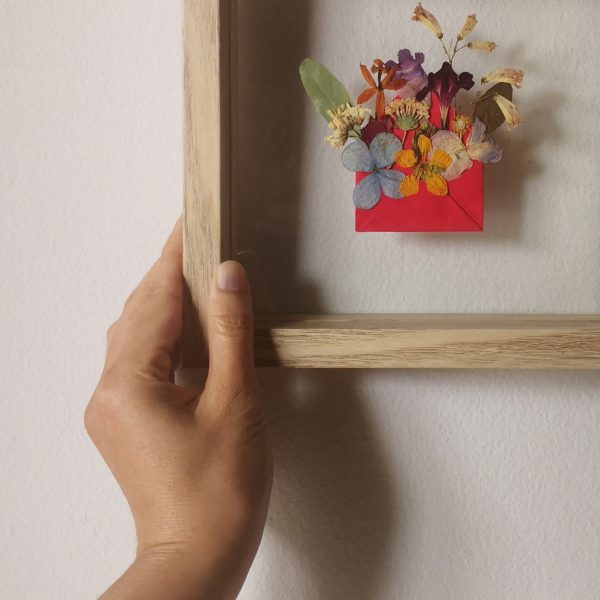 A closeup of Pressed Flower Art Blossom Mail featuring flowers popping out from a red mini envelope and framed in a double glass wooden frame which is being hung on a wall by a left hand