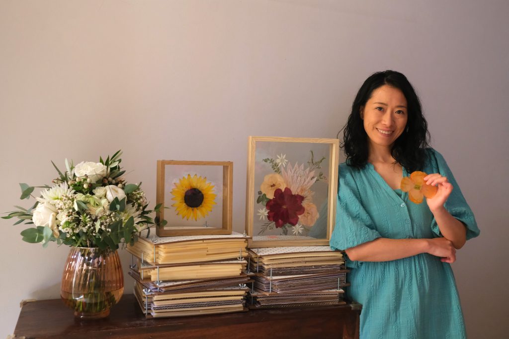 Karin holding a paper-thin poppy leaning on some flower presses, featuring 2 pressed flower art and a bouquet